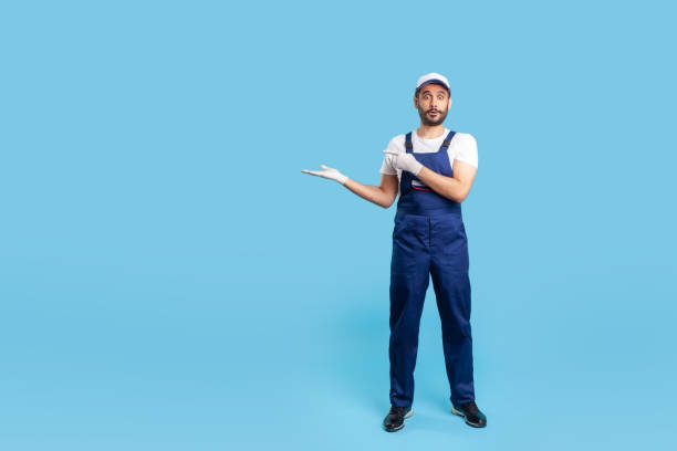 istock Full length surprised workman in blue overalls, cap and protective gloves standing, pointing empty space 1262317678