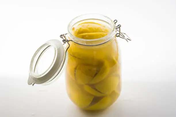 Glass mason jar full of preserved lemons in brine solution shot against bright white background in photographic studio with liquid splashes on the background