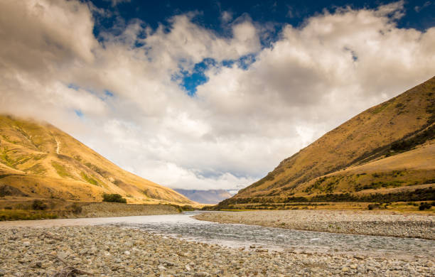 A New Zealand mountain stream in a V shaped valley with dramatic clouds A New Zealand mountain stream, the Ahuriri River, in a V shaped valley with dramatic clouds in the background omarama stock pictures, royalty-free photos & images