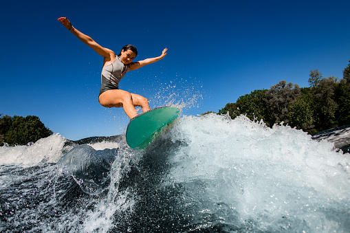 happy woman wakesurfer in gray swimsuit jumps with bright surf board. Clear blue sky and green trees in the background.