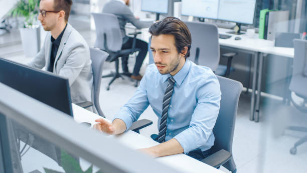 istock Handsome Motivated Office Worker Uses Desktop Computer, Typing on a Keyboard. Confidentand Young Businessman Works in the Big Bright Office with Team of Professionals 1262358991