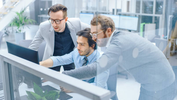 istock In Bright Modern Office: Businessman Sitting and Working at His Desktop Computer, Project Manager and Team Leader Standing Beside Him. They Have Discussion, Find Problem Solution, Pointing at Screen 1262359007