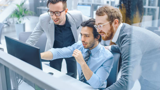 istock In Bright Modern Office: Businessman Sitting and Working at His Desktop Computer, Project Manager and Team Leader Standing Beside Him. They Have Discussion, Find Problem Solution, Pointing at Screen 1262359022