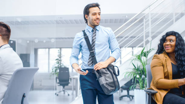 young happy businessman walks into bright modern office, holds smartphone and takes place at his desktop computer. in the background diverse team of professional businesspeople working - vacations women men stability imagens e fotografias de stock