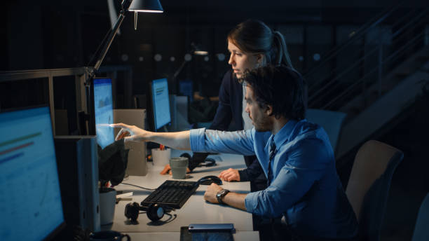 istock Ambitious Businessman Uses Desktop Computer, His Female Project Manager Explains Specific Tasks, Account Handling and Strategic Moves. Professional People Late at Night in Big Corporate Office 1262359154