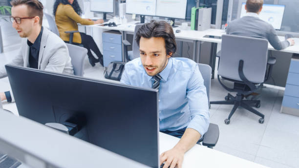 istock Handsome Motivated Office Worker Uses Desktop Computer, Typing on a Keyboard. Confidentand Young Businessman Works in the Big Bright Office with Team of Professionals 1262359168