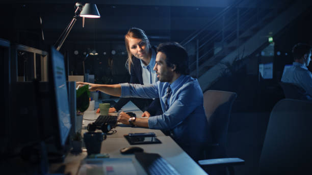 Ambitious and Confident Businessman Uses Desktop Computer, His Female Project Manager Explains Specific Tasks, Account Handling, Strategic Moves. Professional People Late at Night in Corporate Office empresario ambicioso y confiado utiliza computadora de escritorio, su gerente de proyecto femenino explica tareas específicas, manejo de cuentas, movimientos estratégicos. personas profesionales tarde en la noche en la oficina corporativa