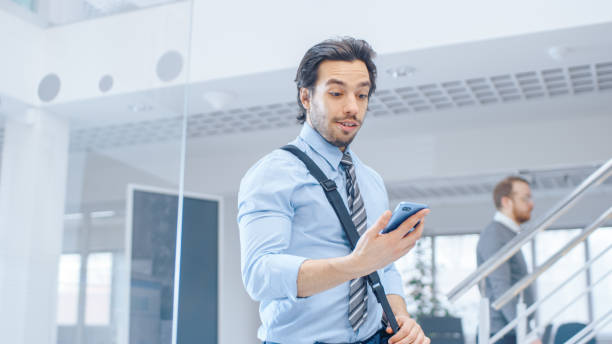 young happy businessman walks into bright modern office, holds smartphone and checks social media. in the background diverse team of professional businesspeople working - vacations women men stability imagens e fotografias de stock