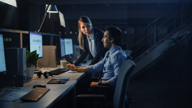 istock Ambitious Businessman Uses Desktop Computer, His Female Project Manager Explains Specific Tasks, Account Handling and Strategic Moves. Professional People Late at Night in Big Corporate Office 1262359230