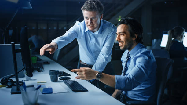istock Ambitious and Confident Businessman Uses Desktop Computer, His Project Manager Explains Specific Tasks, Account Handling and Strategic Moves. Professional People Late at Night in Big Corporate Office 1262359234