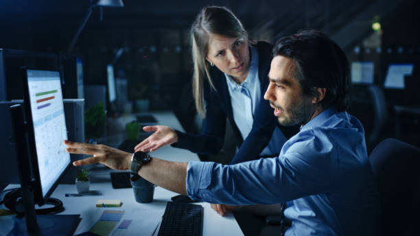 istock Confident Businessman Uses Desktop Computer, His Female Manager Explains Specific Tasks, Account Handling and Strategy. Professional People Discussion and Arguing Late at Night in Corporate Office. 1262359249
