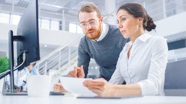 istock In Bright Modern Office: Beautiful Businesswoman Sitting and Working at Her Desktop Computer with Project Manager Standing Beside Her, They Have Discussion, Find Problem Solution 1262359259