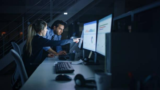 istock Confident Businessman Uses Desktop Computer, Her Male Manager Explains Specific Tasks, Account Handling and Strategy. Professional People Debating, Discussing Issues Late at Night in Corporate Office 1262359267