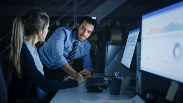 istock Confident Businessman Uses Desktop Computer, Her Male Manager Explains Specific Tasks, Account Handling and Strategy. Professional People Debating, Discussing Issues Late at Night in Corporate Office 1262359303