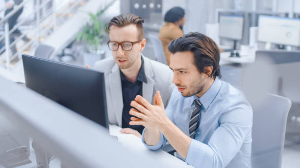 istock In Bright Modern Office: Handsome Office Worker Sitting and Working at His Desktop Computer, Discusses Problem Solution with His Male Project Manager. Diverse Team of Businesspeople Solving Problems 1262359306