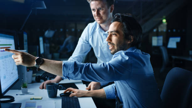istock Ambitious and Confident Businessman Uses Desktop Computer, His Project Manager Explains Specific Tasks, Account Handling and Strategic Moves. Professional People Late at Night in Big Corporate Office 1262359341