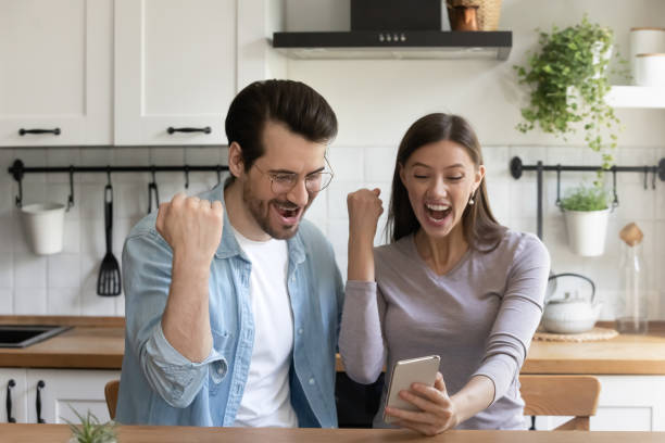 Overjoyed millennial couple triumph win lottery on cell Excited young Caucasian husband and wife sit at kitchen feel euphoric win online lottery on smartphone app, overjoyed millennial couple triumph read unexpected news message or sale deal on cellphone unusual couple stock pictures, royalty-free photos & images
