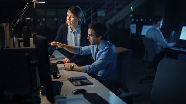 istock Ambitious Businessman Uses Desktop Computer, His Female Project Manager Explains Specific Tasks, Account Handling and Strategic Moves. Professional People Late at Night in Big Corporate Office 1262376862