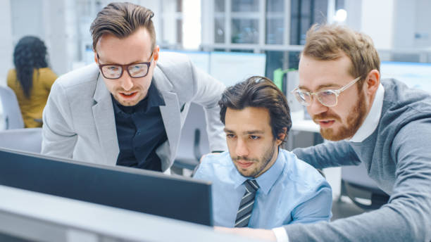 istock In Bright Modern Office: Businessman Sitting and Working at His Desktop Computer, Project Manager and Team Leader Standing Beside Him. They Have Discussion, Find Problem Solution, Pointing at Screen 1262376959