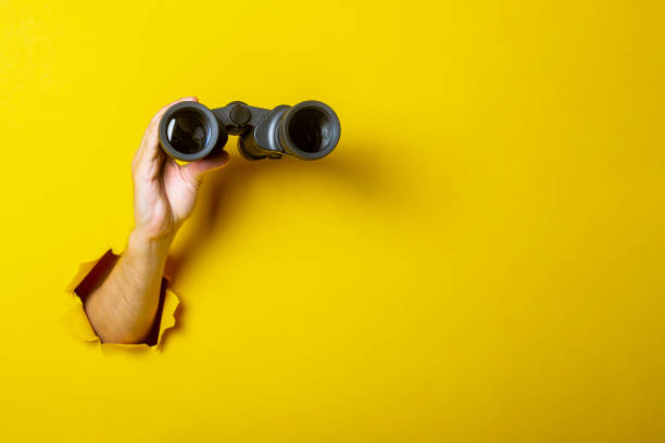 istock Female hand holds black binoculars on a yellow background. Looking through binoculars, journey, find and search concept 1262392393