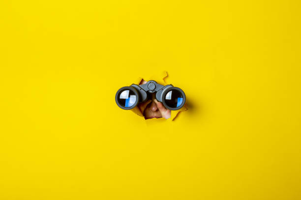 female hand holds black binoculars on a yellow background. journey, find and search concept - finding imagens e fotografias de stock