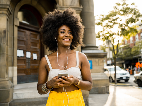 Young African American woman feeling good while listening to music from smart phone with earbuds. Urban exterior in the summer.