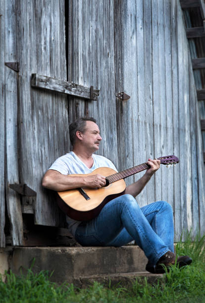 man playing acoustic guitar leaning against barn - musica sertaneja imagens e fotografias de stock