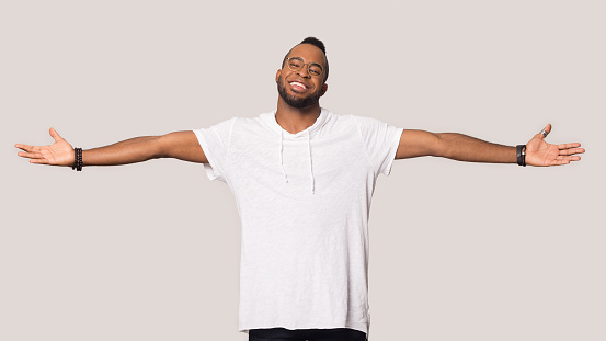 Happy african American man in casual clothes and glasses stretch hands wide open ready for hug, smiling black male look at camera isolated on grey studio background show big sale or great offer