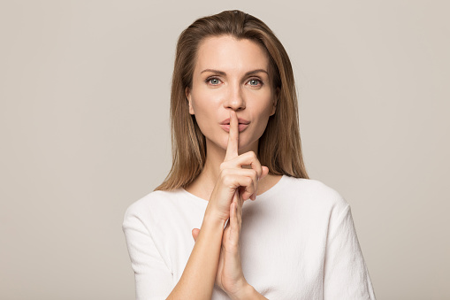 Beautiful young Caucasian woman isolated on grey studio background hold finger at lips ask be silent quiet, millennial female look at camera tell share mystery, announce secret sale or discount