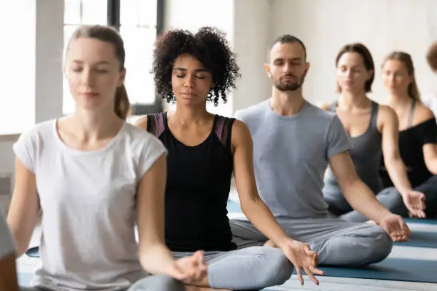 Focus on young beautiful peaceful african american woman sitting in row with mindful diverse people in lotus padmasana pose with folded in mudra sign hands and closed eyes, meditating at yoga class.