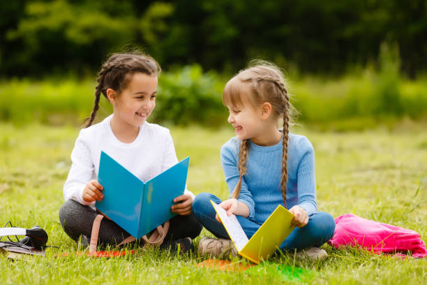 istock two cute multicultural schoolgirls sitting on lawn under tree and reading book together 1262415519