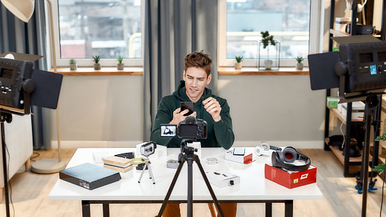 Male technology blogger recording video blog or vlog review of new smartphone, showing it on camera at home studio. Blogging, Work from Home concept. Selective focus. Web Banner
