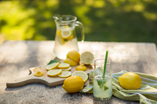 High angle view of a jar and glass of homemade lemon juice prepared in the table of the backyard to enjoy a sunny spring afternoon. The cutting board with slices of lemon is visible.