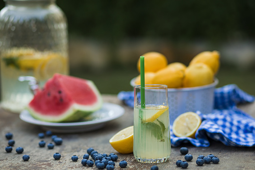 Close up of a glass of homemade lemonade with mint, placed on the table and surrounded with piece of watermelon, more lemons and scatered blueberries.