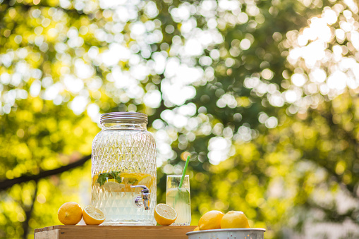 Low angle view of drinks station with lemonade glass, big drinking jar and lemons placed in an open area for people to enjoy an organic natural drink during summer.