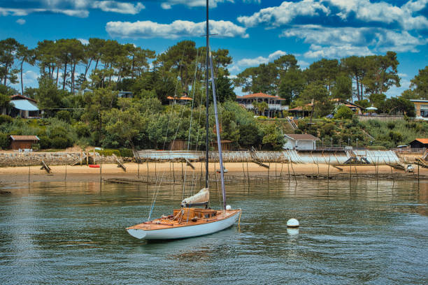 La Vigne, France - July 10, 2020. View of a wooden sailboat hanging from its anchorage in the Bassin d'Arcachon under a blue and cloudy sky. In the foreground, the wooden sailboat is visible. In the background, the coast is visible in front of the sky and its clouds.