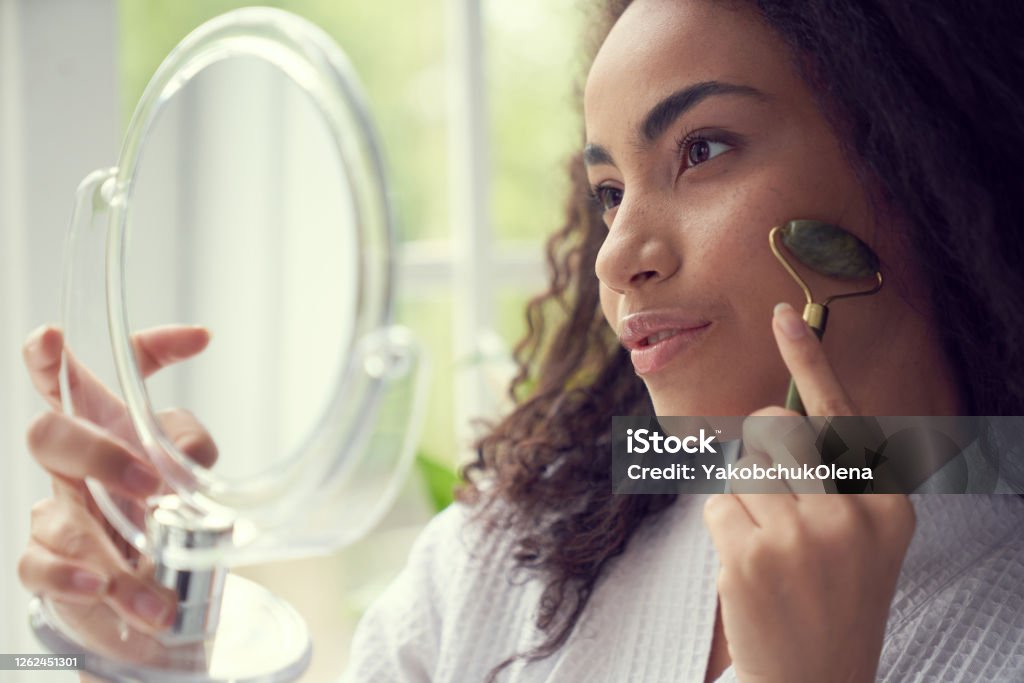 Young woman using jade roller at home Curly attractive female is holding mirror and looking into it while doing face massage indoors Jade Roller Stock Photo