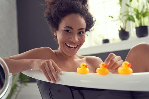 Portrait of merry young curly female having bath and playing with rubber ducks in morning