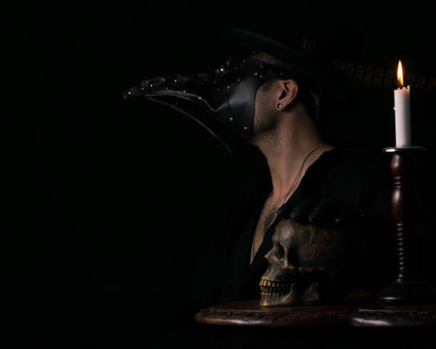 Modern Plague Doctor Medieval inspired image of a young man wearing a plague doctor mask, hand on skull, illuminated by candlelight. black plague doctor stock pictures, royalty-free photos & images