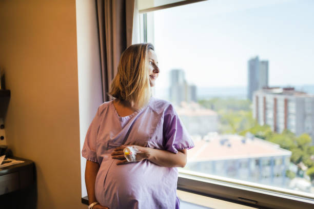 молодая беременная женщина касаясь желудка в ожидании travail к окну в больничной палате - delivery room стоковые фото и изображения