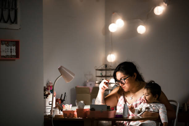 Mother Working From Home at Desk While Holding Baby Daughter Authentic image of a mother working from home at her desk while holding her baby daughter. Shot in the late hours of the evening, the lighting creates the late night mood. working late photos stock pictures, royalty-free photos & images