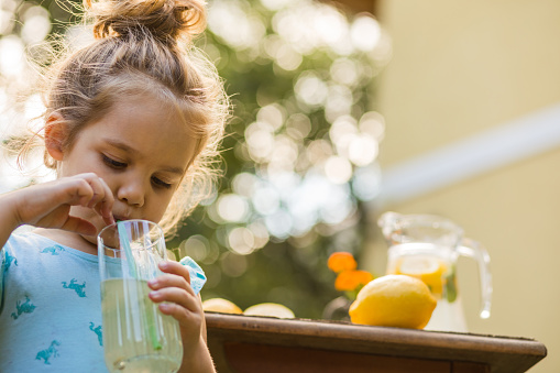 Low angle view of thirsty little girl using a straw to drink a refreshing homemade lemonade in the backyard of her house.