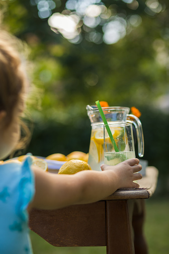Rear view of little girl taking a glass of organic fresh lemonade from the backyard table to get refreshed during summer.