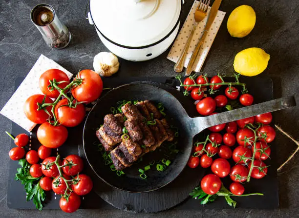 Mediterranean Meat rolls such as Cevapcici, Mititei, served in an iron pan on a dark table with a lot of fresh and ripe tomatoes - ready to eat - top view