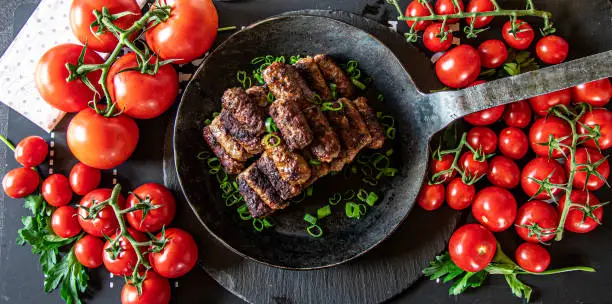 Mediterranean Meat rolls such as Cevapcici, Mititei, served in an iron pan on a dark table with a lot of fresh and ripe tomatoes - ready to eat - top view