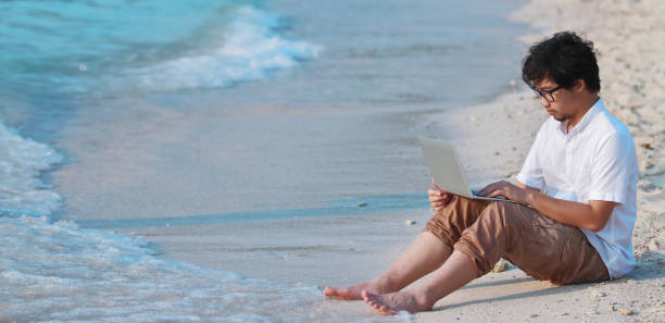 istock Panoramic view of relaxed young Asian man with computer laptop on the sandy beach in summer time. Summer vacations and lifestyle concept. 1262587343