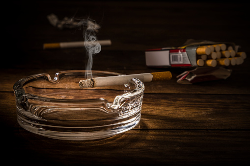 Front view of a burning and smoking cigarette on a crystal ashtray. The ashtray is at the lower left corner and at the background is a defocused cigarette pack. Predominant colors are brown and black. Low key DSLR photo taken with Canon EOS 6D Mark II and Canon EF 24-105 mm f/4L