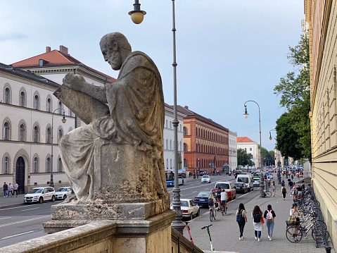 Munich, Germany - July, 21 - 2020: Ludwigstraße, seen from the entrance of the Bayerische Staatsbibliothek (Bavarian state library). It was built in the 19th century. The statue shows Thucydides, the founder of scientific historiography. The planning of the library was entrusted to the architect Friedrich von Gärtner and realized between 1832 and 1843. The elongated building on Ludwigstrasse, which surrounds two inner courtyards, is the largest bright brick building in Germany with a length of 152 meters, depth of 78 meters and a height of 24 meters.