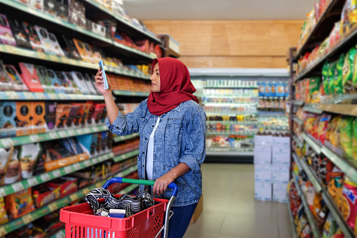 Photo of a Southeast Asian muslim woman reading food label of a chocolate bar during her weekly grocery shopping