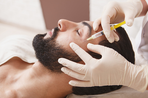 Bearded man getting anti-wrinkle face filler treatment by beautician